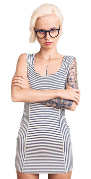 Young blonde woman with tattoo wearing casual clothes and glasses skeptic and nervous, disapproving expression on face with crossed arms. negative person.