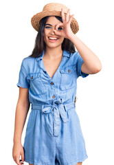 Fototapeta premium Brunette teenager girl wearing summer hat doing ok gesture with hand smiling, eye looking through fingers with happy face.