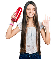 Young brunette girl with long hair holding shampoo bottle doing ok sign with fingers, smiling...