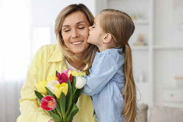 Little daughter kissing and congratulating her mom with Mother`s Day at home. Woman holding bouquet of beautiful tulips