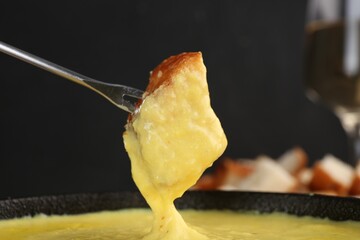 Dipping piece of bread into fondue pot with tasty melted cheese against dark gray background,...