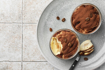 Delicious tiramisu in glasses, spoon and coffee beans on light tiled table, top view. Space for text