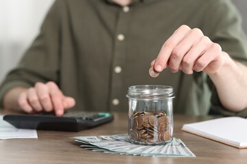 Fototapeta na wymiar Financial savings. Man putting coin into glass jar while using calculator at wooden table, closeup