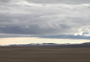 long view of farmers feild with snow topped mountains, white sky, dark cloud swirls formation - Powered by Adobe