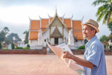 smiling happy asian senior male tourist wearing hat,holding a paper map,standing in front of temple,elderly man religion travel destination in south east asia,Thailand