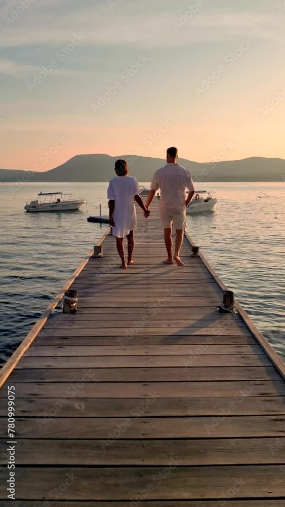 Wall mural A man and a woman standing on a dock by the ocean, holding hands admiring the horizon. Their gestures express happiness and contentment as they enjoy the leisure of being in nature, Samaesarn Thailand