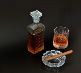 A glass ashtray with a cigar, a crystal decanter and a glass of whiskey on a black background.