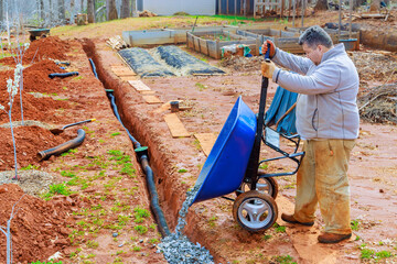 Using wheelbarrow, worker fills over drainage pipes with crushed stone
