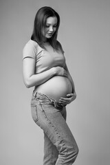 Black and white portrait of young pretty pregnant woman in t-shirt and jeans on gray background.