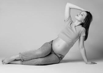 Black and white photo of young pretty pregnant woman in t-shirt and jeans sit on the floor on gray background. Female with belly exposed. - 781014080