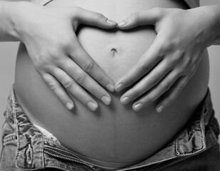 Black and white photo of  female on 6th month of pregnancy. Woman touching with hands her naked belly and shows heart sign. - 781014081