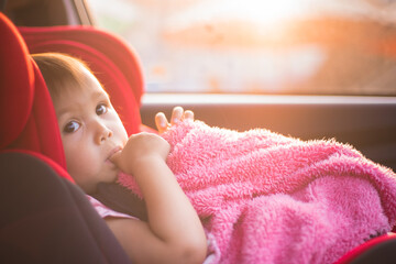 Asian Toddler Girl seats peacefully in her red carseat. She is holding her pink blanket while...