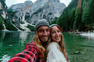 Fototapeta na wymiar A young couple taking a selfie photo on Lake Braies in Dolomites. The man is wearing a red flannel shirt and grey beanie, the woman wears a white sweater with gray fur hat smiling at the camera