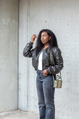 In this contemporary urban portrait, a stylish young woman leans casually against a concrete wall. She's fashion-forward in a sleek leather jacket and grey jeans, her hair falling in natural waves