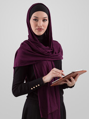 Beautiful and modern young Arabic businesswoman in hijab using a tablet computer while posing on light background and smiling at the camera. Business diversity concept, Muslim lady. - 781034259