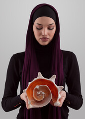 Pretty stylish Muslim woman wearing hijab and holding a seashell and dreams closed eyes. Golden...