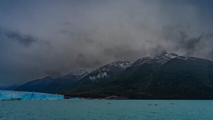Canoes with tourists sail along the turquoise glacial lake to the glacier. A wall of blue ice rises...