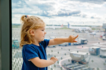 Fototapeta na wymiar Cute little toddler girl at the airport, traveling. Happy healthy child waiting near window and watching airplanes. Family going on summer vacations by plane.