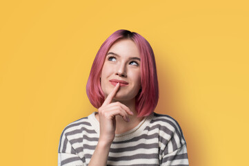 Portrait ad image - mysteriously grinning smile look up thinking gladly pink hair woman wear grey striped sweater jumper, hold finger over her she lips, isolated yellow studio wall background.