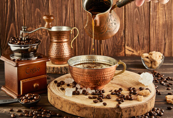 Pouring coffee from coffee maker into copper cup, an antique coffee grinder and copper milk jug on wooden background