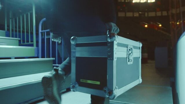 Man carries weighty suitcase filled with instruments. Worker walks along long rows of seats in concert hall holding suitcase in hand