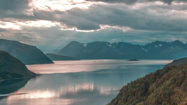 Rays of the setting sun shine through the storm clouds above the serene fjord in a timelapse video.