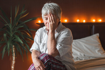 Sad unhappy senior woman seated on bed in bedroom
