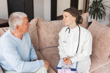 Female professional doctor showing medical test result explaining prescription using clipboard visiting senior elderly old man patient at home sitting on sofa - 781102676