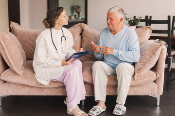 Female professional doctor showing medical test result explaining prescription using clipboard visiting senior elderly old man patient at home sitting on sofa - 781102683