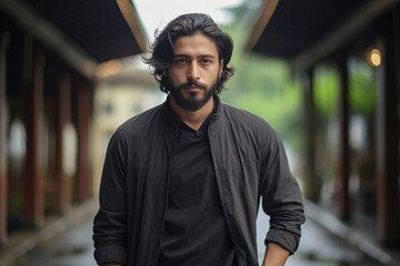 A man with a beard and long hair stands in front of a building