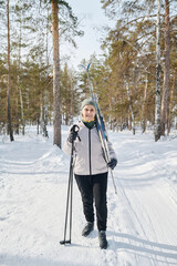 Vertical long shot of cheerful Caucasian senior woman walking along forest park carrying skis and poles on winter day, copy space
