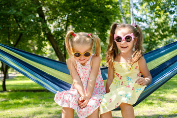 Two cute and playful five-year-old sisters wearing matching colorful sunglasses, happily relaxing side by side in a comfortable hammock at the park on a bright and sunny day.