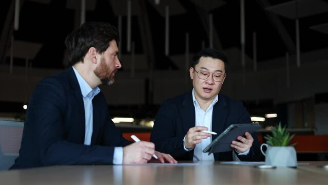 Serious asian sales representative showing new product on tablet to perspective buyer for implementation. Progressive developer explaining all pros and cons of application for corporate interaction.