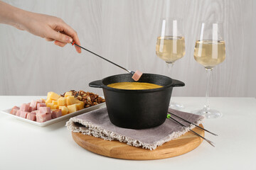 Dipping piece of ham into fondue pot with tasty melted cheese at white table, closeup