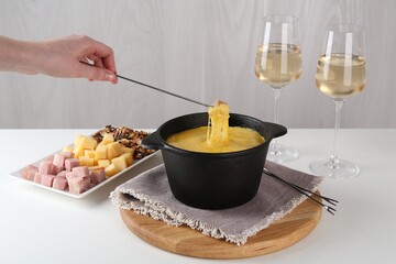 Dipping piece of ham into fondue pot with tasty melted cheese at white table, closeup