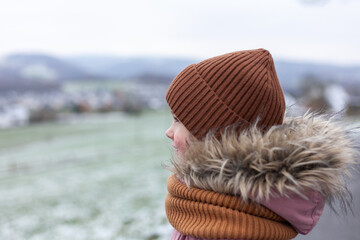 A little girl in a warm hat and scarf stands on the background of a winter landscape.
