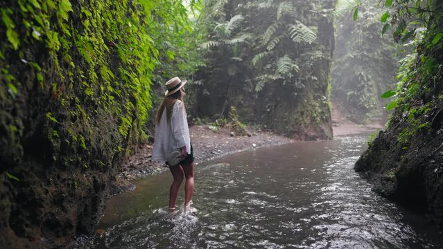 Woman tourist in straw hat taking picture recording video on camera of huge waterfall among tropical mountains on island. Travelling in wild nature admiring views. Travel tourism, adventure concept.