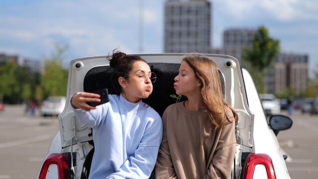 Beautiful happy teen girl sitting in car trunk with pretty mom taking selfie photos on smartphone. Caucasian woman with daughter or sister making funny face expression posing to cellphone