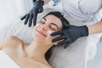 A cosmetologist applies cream to the face of a woman lying on a couch. Facial. Facial skin care...