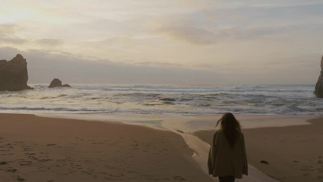 Girl walking towards the ocean on a remot sand beach surrounded by cliffs at sunset. Drone pedestal shot