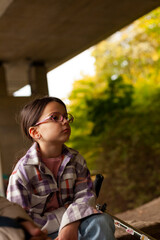 A tired little girl in a plaid shirt and glasses sits sadly under a bridge next to a scooter