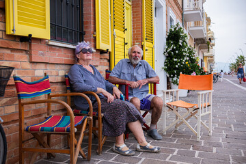 An elderly couple sits on a colorful chair near a brick wall of a house with yellow shutters