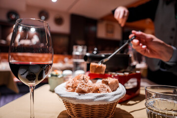 Intimate dining scene with red wine, bread basket, fondue pot, person dipping food. Cozy ambiance, warm lighting. Possible location Europe or North America.