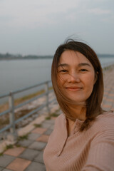Vertical photo of asian Thai woman use mobile taking selfie while relaxing and standing alone along the river, sunset time, happy life traveling concept. 