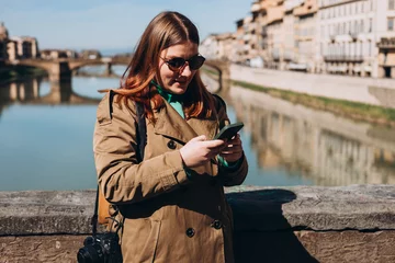 Photo sur Plexiglas Ponte Vecchio Happy 30s girl using smartphone on famous Old bridge in Florence. Stylish woman visiting Italian landmarks. Concept of travel, tourism and vacation in city. Woman walking on city street checks phone.