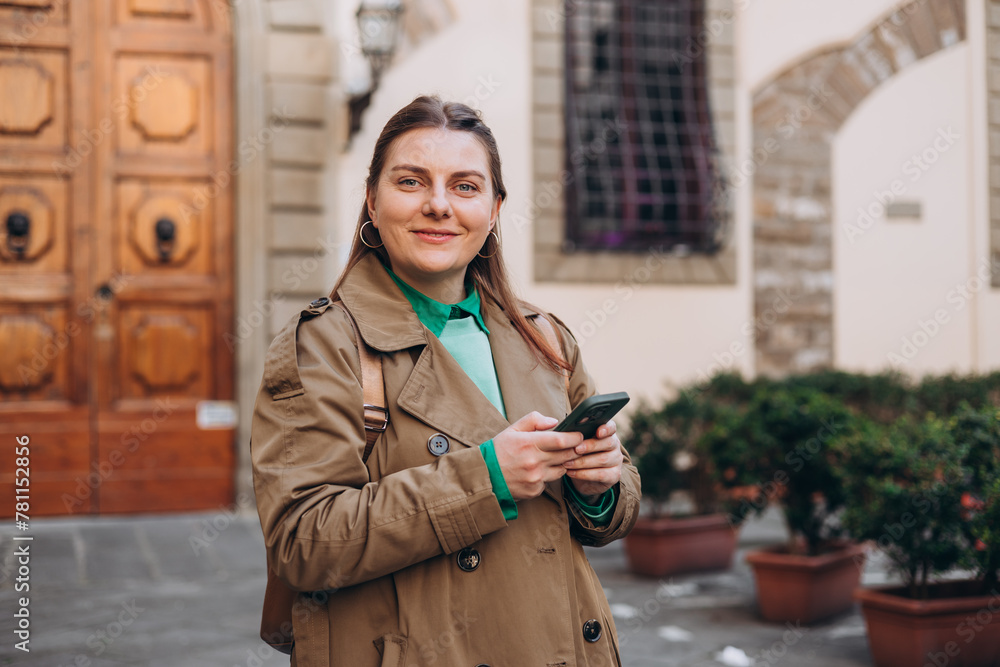 Wall mural Happy cheerful young woman with backpack walking on city street checks her smartphone. Portrait of beautiful 30s girl using smartphone outdoors. Spring time. Concept of travel, technology and vacation