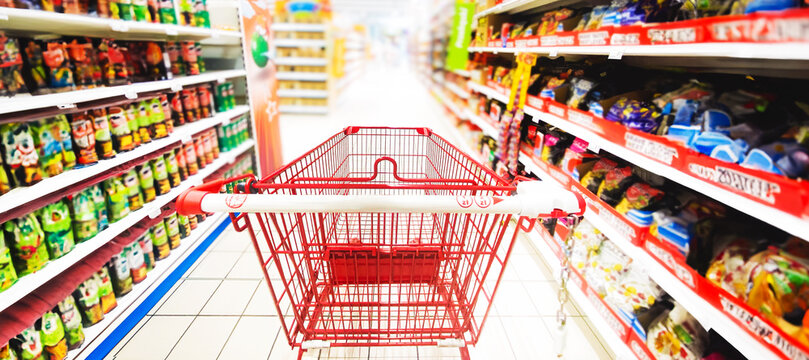 Pusty koszyk, w&oacute;zek sklepowy w supermarkecie, rozmazane tło, wnętrze sklepu, supermarketu, w&oacute;zek na zakupy, p&oacute;łki sklepowe z produktami. Hipermarket. Baner, miejsce na tekst.