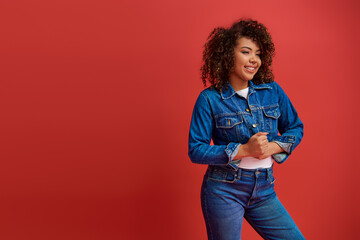 joyous elegant african american woman in stylish denim outfit looking away on red backdrop