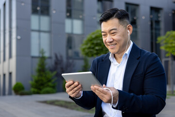 Close-up photo of a smiling young Asian man sitting outside in a suit and headphones, holding a tablet and talking on a video call
