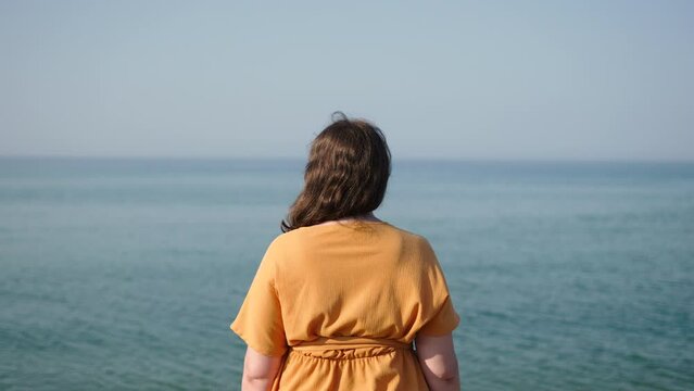 Unrecognizable Caucasian woman stands on seashore and looks into horizon. Sad lady in depression and despair in good weather reflects on beach in orange dress in sunny calm weather.